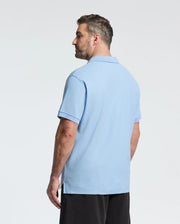 A bearded man with short hair, sporting Psycho Bunny's Big and Tall Classic Pique Polo (B9K001CRPC) in light blue and black pants, stands against a plain white background, seen from the side with a slight turn.