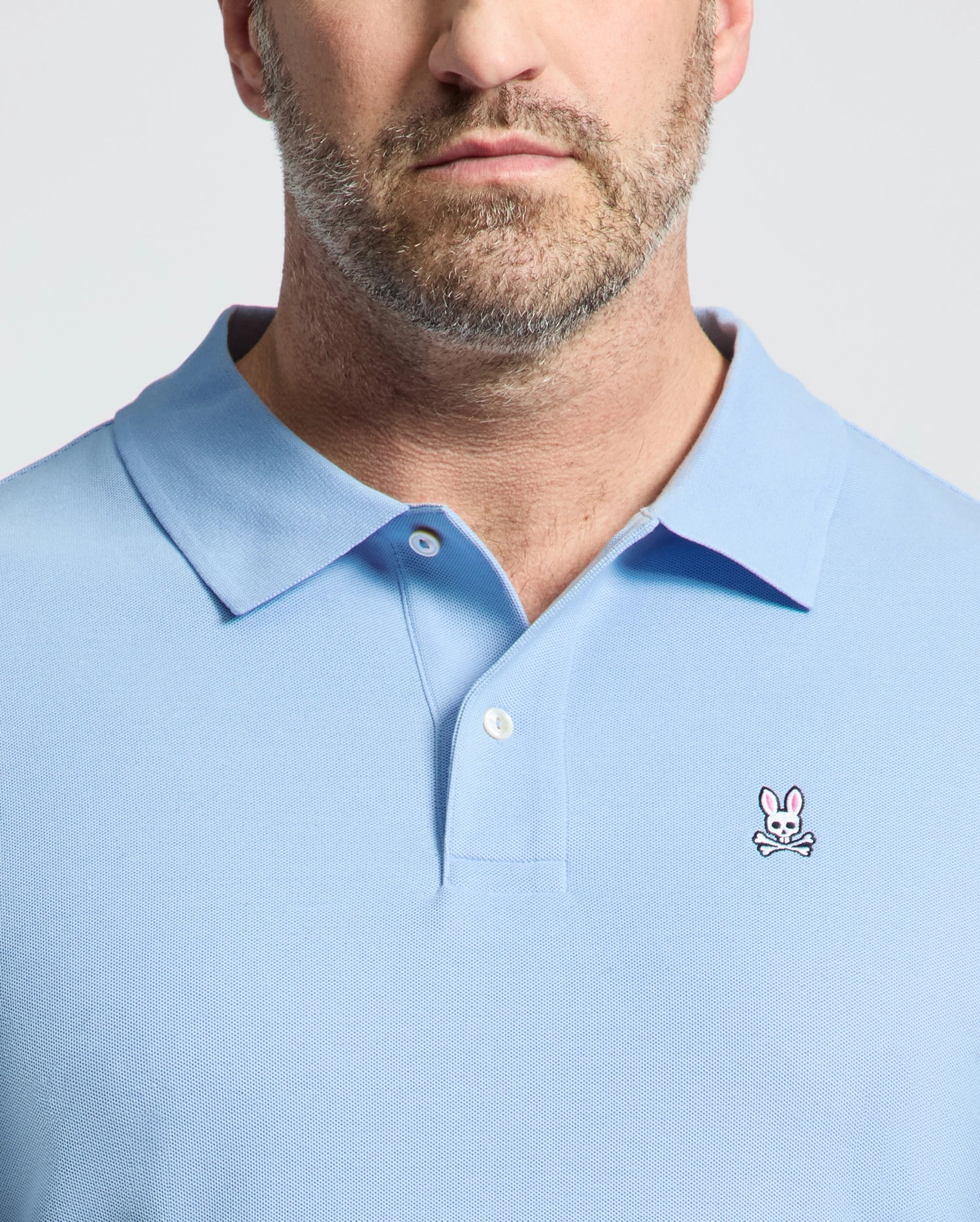 A bearded person is wearing a light blue Psycho Bunny BIG AND TALL CLASSIC PIQUE POLO, featuring a small pink bunny with crossbones logo on the chest, with focus on the upper torso and this stylish symbol.