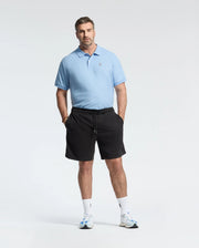 A man in a Psycho Bunny BIG AND TALL CLASSIC PIQUE POLO - B9K001CRPC stands against a white background, wearing black shorts and white sneakers with blue accents. He effortlessly sports the light blue polo, hands in pockets, gazing at the camera.