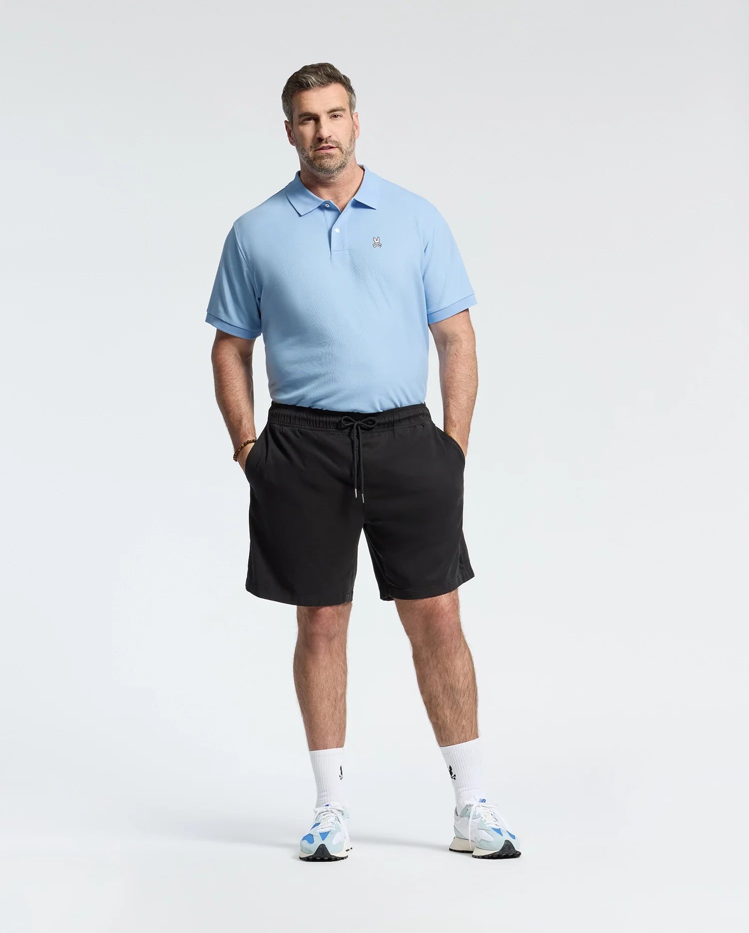 A man in a Psycho Bunny BIG AND TALL CLASSIC PIQUE POLO - B9K001CRPC stands against a white background, wearing black shorts and white sneakers with blue accents. He effortlessly sports the light blue polo, hands in pockets, gazing at the camera.