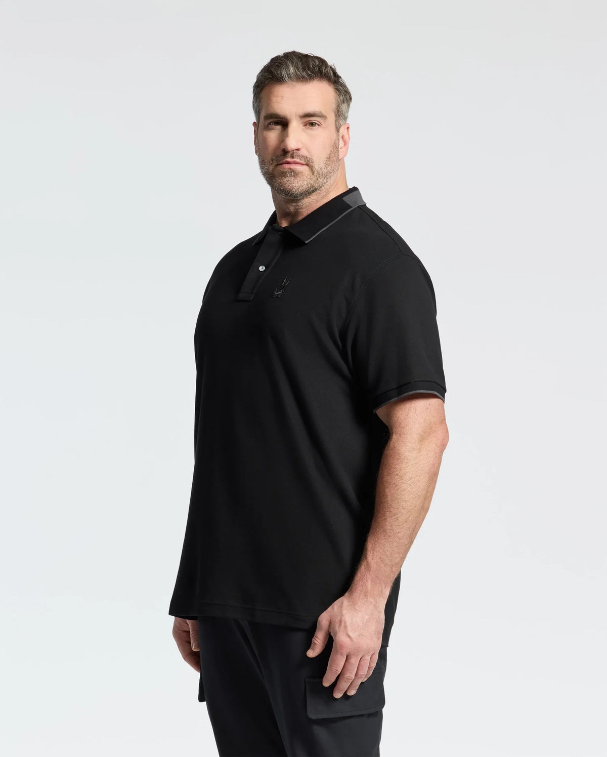A person with short hair and a beard is wearing a Psycho Bunny MENS BIG AND TALL NEW CASTLE PIQUE POLO - B9K487F200, made of 100% Pima cotton, paired with dark pants. They stand confidently against a plain white background, showcasing their attire's luxury finishes.