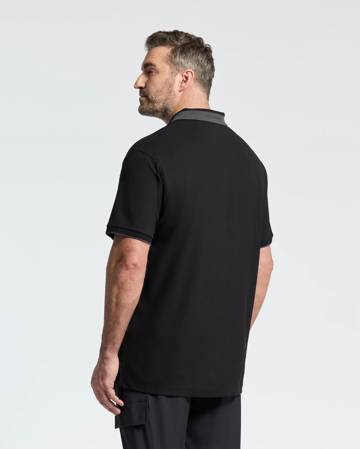 A bearded man with short hair stands looking sideways in a Psycho Bunny MENS BIG AND TALL NEW CASTLE PIQUE POLO, featuring luxury finishes and gray trim, paired with dark pants. The light-colored background highlights his stylish yet understated appearance.