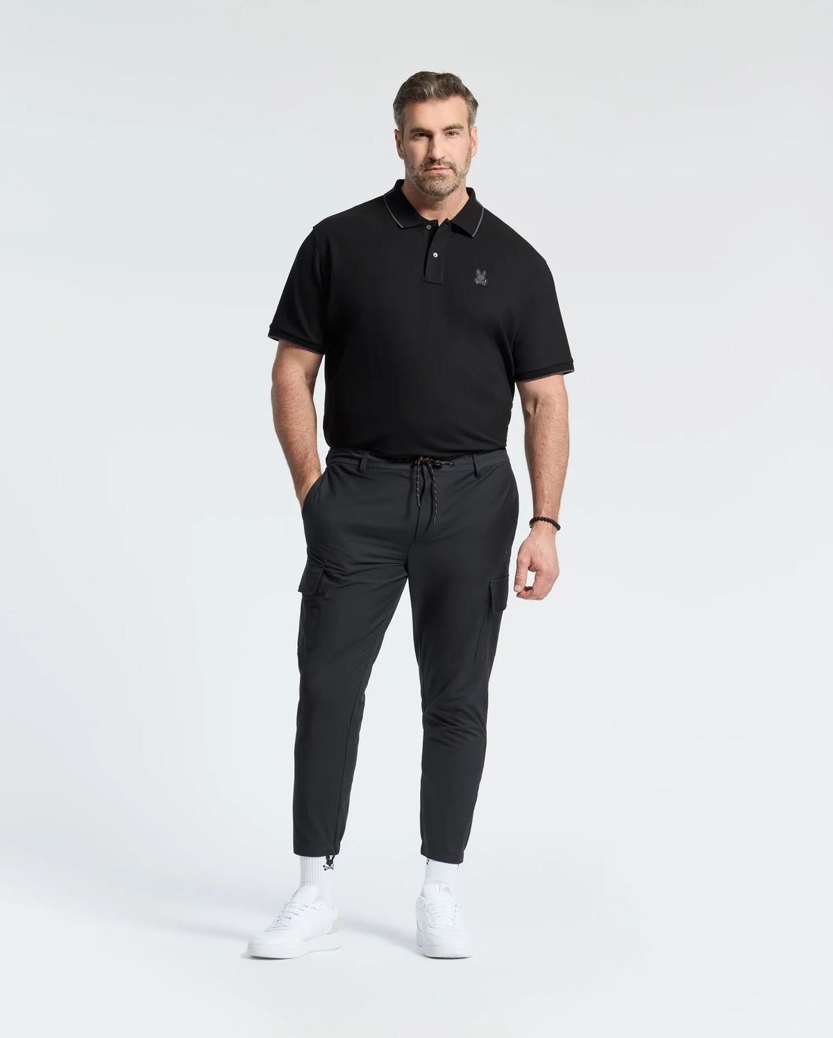 A man with a neat beard stands against a white background wearing a Psycho Bunny MENS BIG AND TALL NEW CASTLE PIQUE POLO in black, black cargo pants, and white sneakers. His hands are in his pockets.
