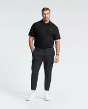 A man with a neat beard stands against a white background wearing a Psycho Bunny MENS BIG AND TALL NEW CASTLE PIQUE POLO in black, black cargo pants, and white sneakers. His hands are in his pockets.