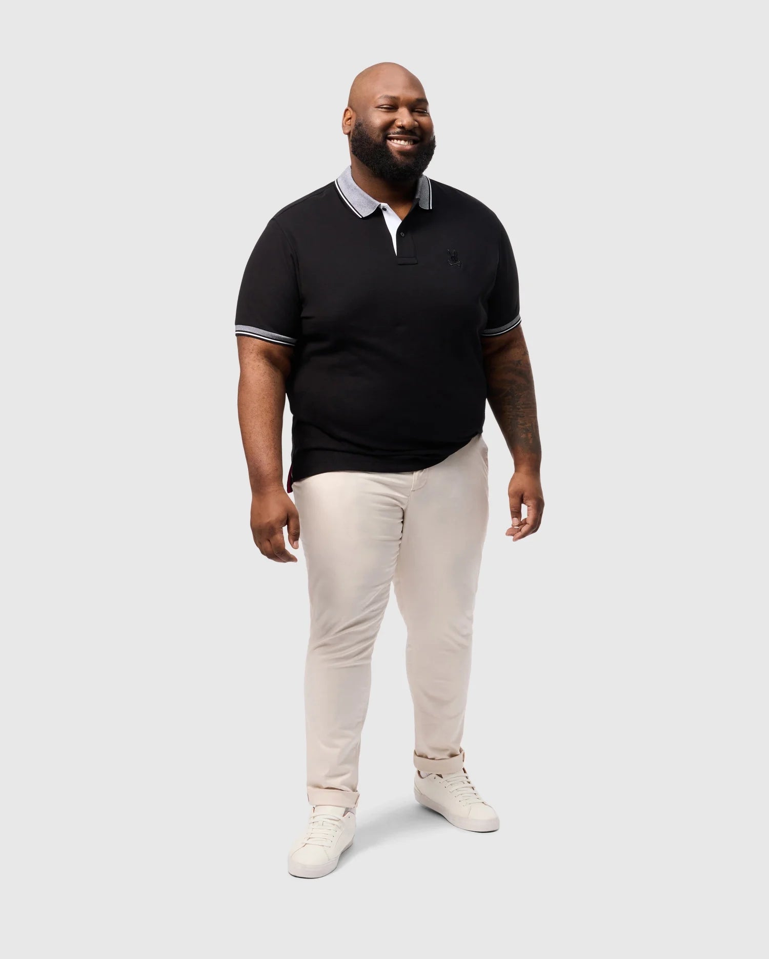 A man smiles in a Psycho Bunny BIG AND TALL MALTON PIQUE POLO SHIRT - B9K950D200, featuring black Pima cotton with white trim. He's paired it with beige pants and white sneakers, standing against a light gray backdrop, reflecting the brand's signature casual elegance.