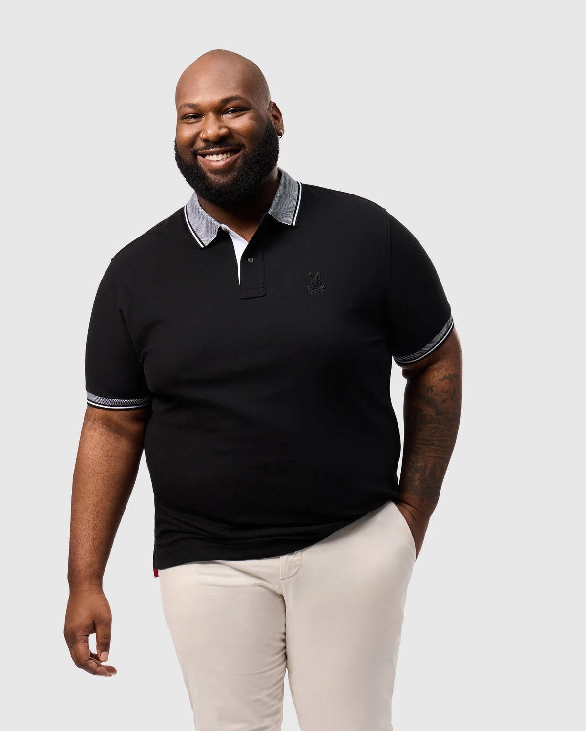A bearded person smiles against a gray background, wearing a Psycho Bunny BIG AND TALL MALTON PIQUE POLO SHIRT with a contrasting collar and light pants. One arm is relaxed at their side, radiating friendliness.