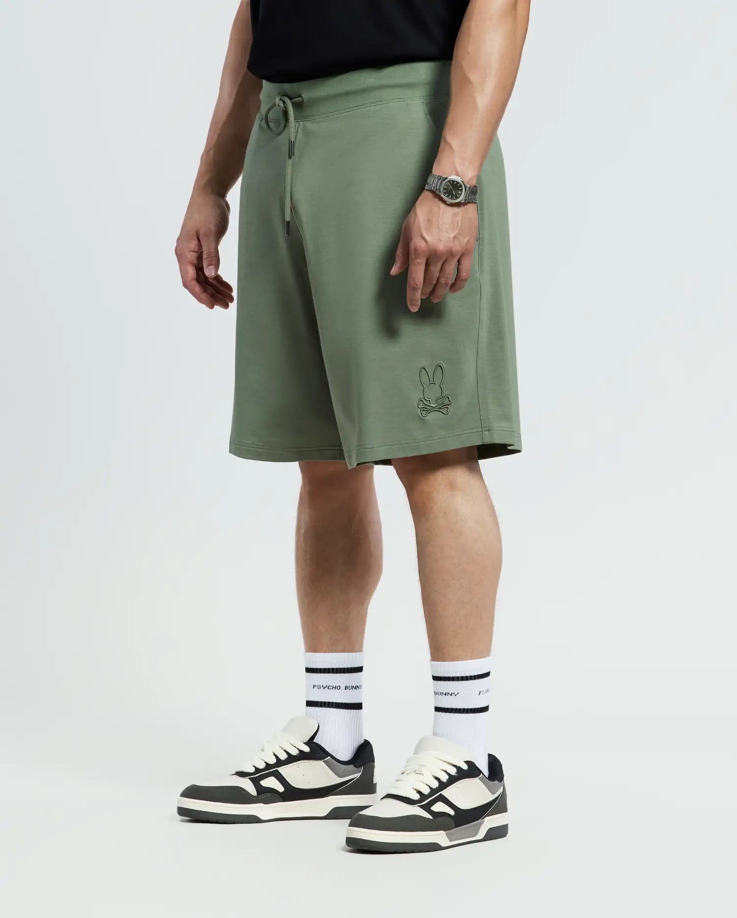 A person is wearing olive Psycho Bunny Big and Tall Levanzo French Terry Sweatshorts, a black T-shirt, black and white sneakers, and white socks. A watch adorns their wrist against a neutral background.