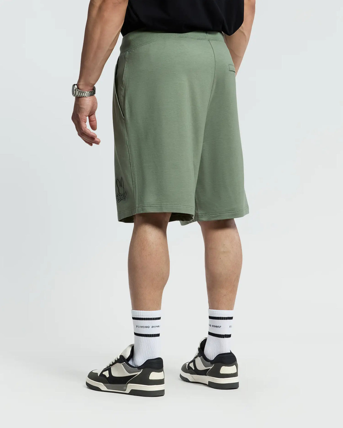 A person in Psycho Bunny's green Levanzo French Terry Sweatshorts, a black T-shirt, white socks, and black sneakers stands with their back to the camera. A watch is on the left wrist against a plain white background.
