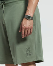 Close-up of a person wearing Psycho Bunny's green Levanzo French Terry Sweatshorts, featuring an embroidered bunny skull logo near the hem. Their hand, with a matching green dial wristwatch, rests by the pocket. The shorts provide comfort and style against a plain backdrop.