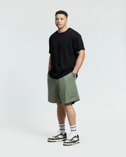 Against a plain backdrop, someone showcases effortless style wearing a black t-shirt with olive green Psycho Bunny Levanzo French Terry Sweatshorts. Paired with white socks and black-and-white sneakers, they stand facing forward with hands in pockets.
