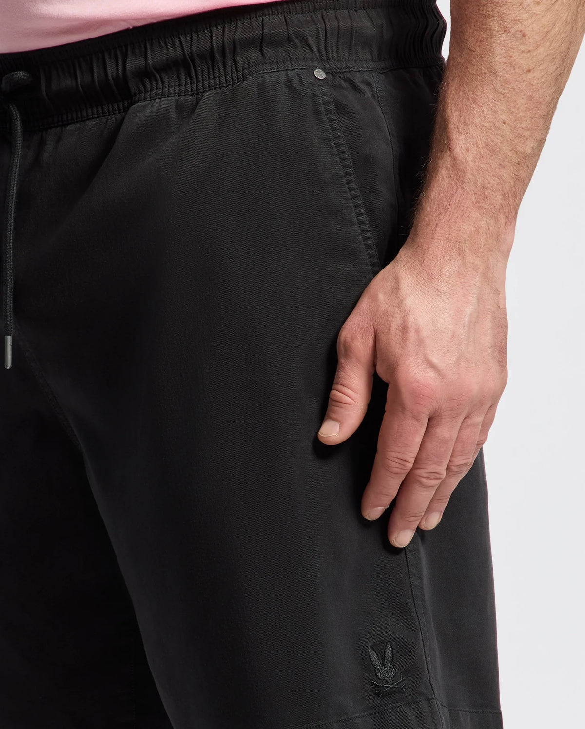 A person wearing Psycho Bunny's Mens Big and Tall Willis Stretch Tencel Shorts, featuring a subtle logo near the pocket, stands with their right hand on their thigh. The elastic waistband with a drawstring provides comfort, while the stretch Tencel fabric ensures ease of movement; a pink shirt peeks out at the top.