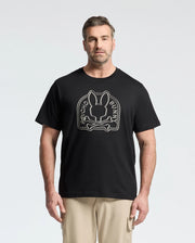 A man in a black MENS BIG AND TALL MELBOURNE GRAPHIC TEE - B9U410F200 by Psycho Bunny, made from eco-friendly Peruvian Pima cotton, features a white outlined "Psycho Bunny" logo with bunny and crossbones. He is against a plain white background, wearing beige pants.