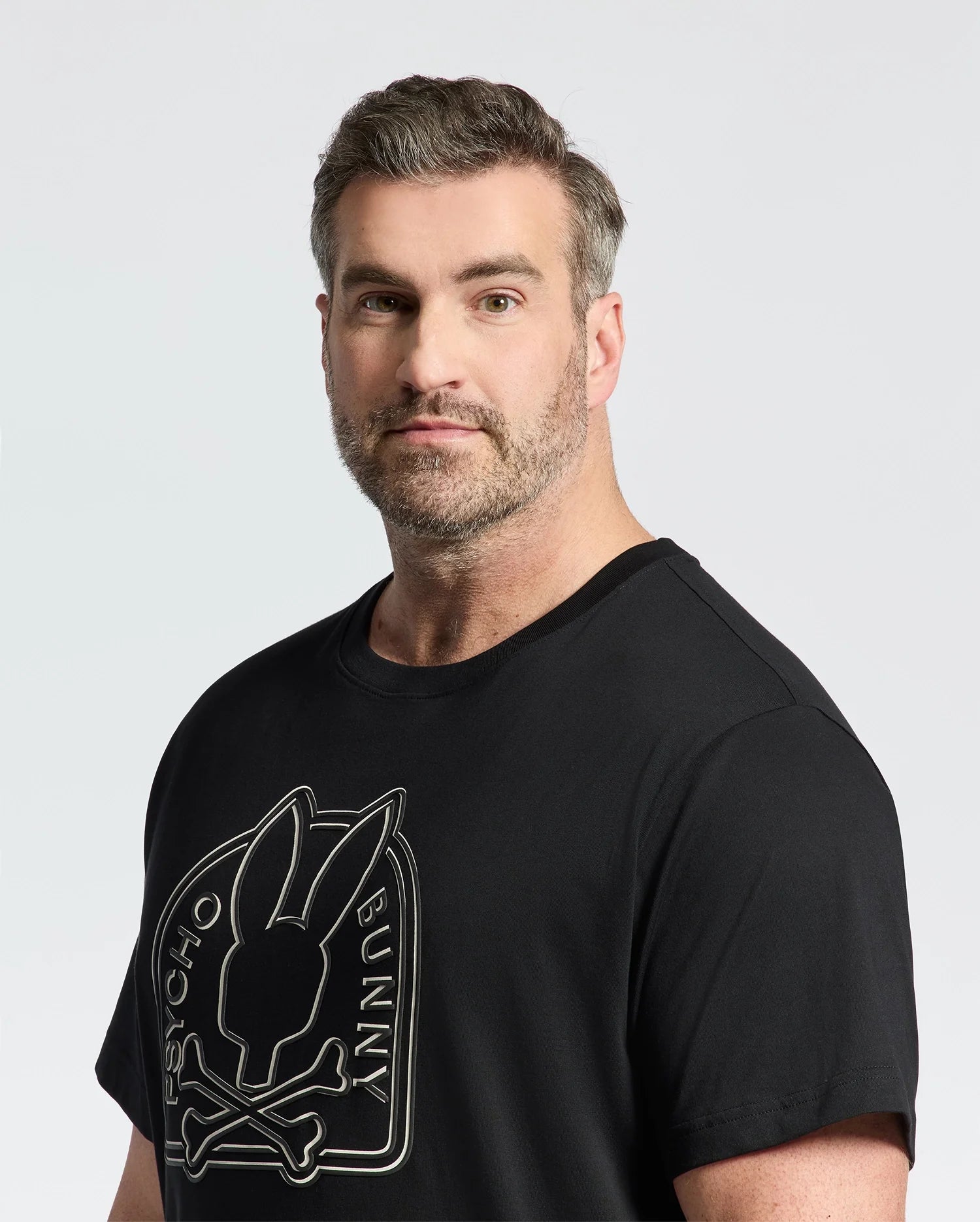 A bearded man with short hair shows off a serious look in the eco-friendly black Psycho Bunny Men's Big and Tall Melbourne Graphic Tee, featuring a bunny and crossbones design. The premium feel of its plain white background is crafted from Peruvian Pima cotton for ultimate comfort.