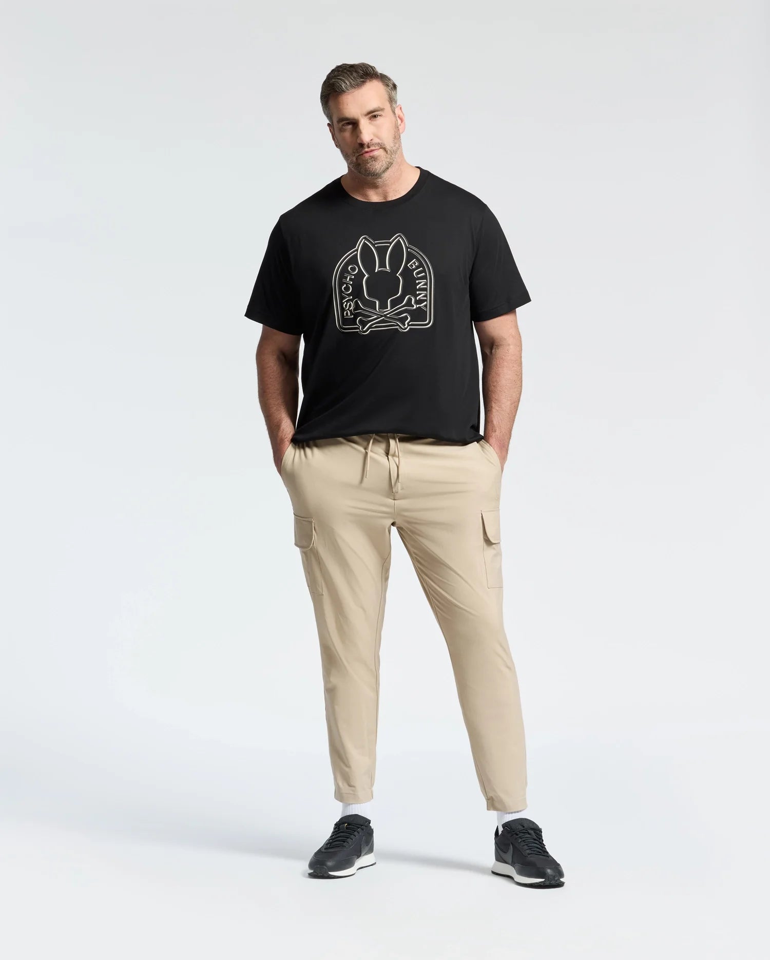 A man wears a black Psycho Bunny Big and Tall Melbourne Graphic Tee (B9U410F200), made from eco-friendly Peruvian Pima cotton, along with beige cargo pants. Standing against a plain background, he has his hands in his pockets and a neutral expression.