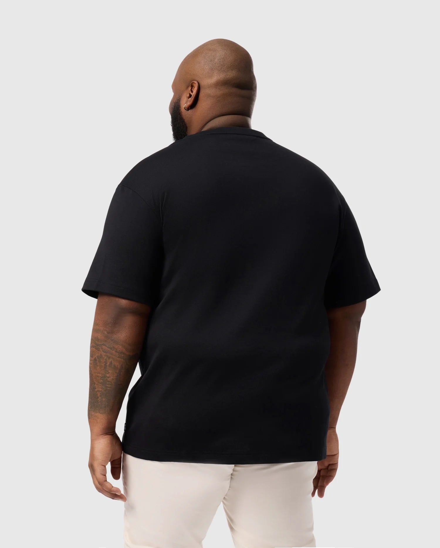 A bald man with a tattoo on his left arm faces away, wearing a Psycho Bunny BIG AND TALL DUKE OVERSIZED TEE (B9U742D200), crafted from heavyweight Pima cotton. His black tee is paired with light-colored pants against a plain, light gray background.