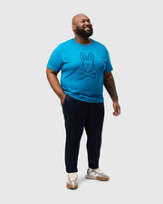 A person in a Psycho Bunny BIG AND TALL MALTON GRAPHIC TEE - B9U795D200 is smiling and standing against a plain background, paired with dark pants and sneakers, embodying the effortless blend of style and detail typical of Peruvian craftsmanship.