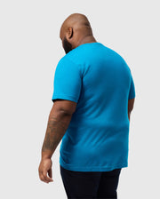 A tattooed person faces away, wearing a Psycho Bunny BIG AND TALL MALTON GRAPHIC TEE (B9U795D200) in blue Pima cotton with dark pants against a plain gray background.