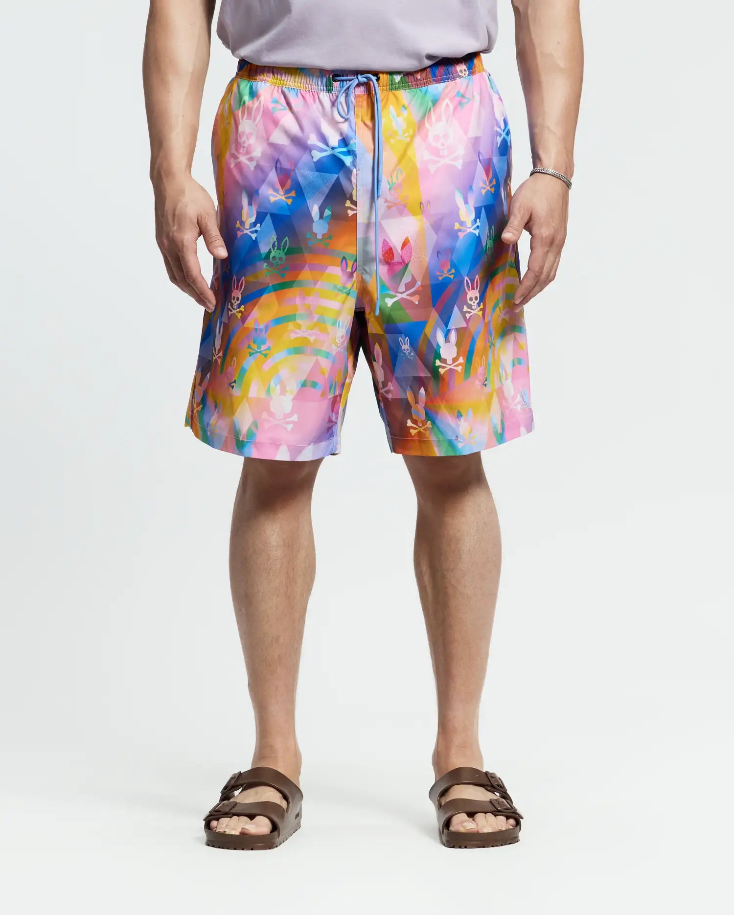 Wearing Psycho Bunny's MENS BIG AND TALL MILOS SWIM TRUNK - B9W446F200 with a bright abstract floral design, the person pairs it with a gray shirt and brown sandals, standing against a plain white backdrop.