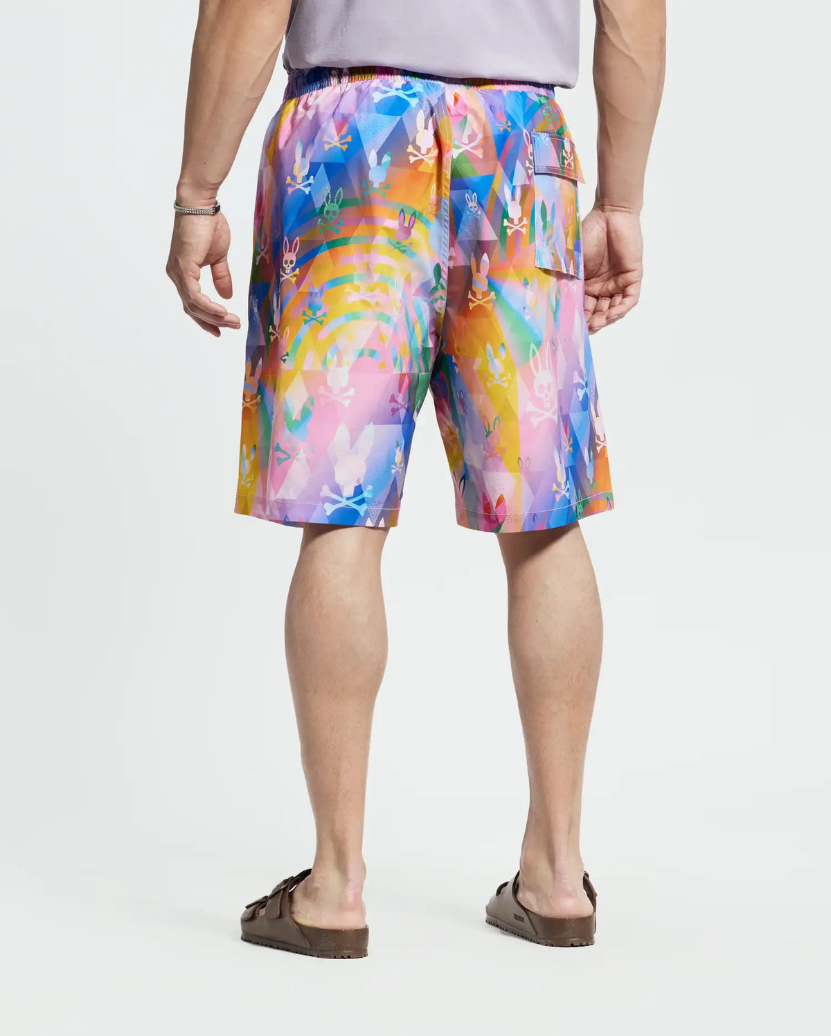 A person is shown from behind wearing a short-sleeved gray shirt, Psycho Bunny Big and Tall Milos Swim Trunk with a colorful abstract pattern of pink, blue, orange, and yellow in quick-dry fabric, and brown sandals.