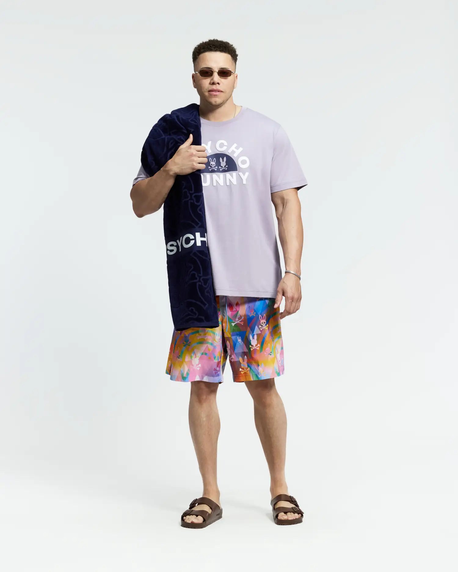 A man wearing sunglasses showcases a "Psycho Bunny" light purple T-shirt and colorful Mens Big and Tall Milos Swim Trunk, made from quick-dry fabric. He completes the look with brown sandals and a dark blue towel over his shoulder against a plain white background.