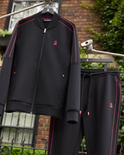 A black Psycho Bunny MENS TRAVIS TRACK JACKET - B6J142D200 and matching jogger pants with pink accents hang on a clothesline. The set features a small pink embroidered bunny logo on the chest of the jacket and hip of the pants, along with invisible zip pockets for added convenience. A brick building with green foliage is blurred in the background.