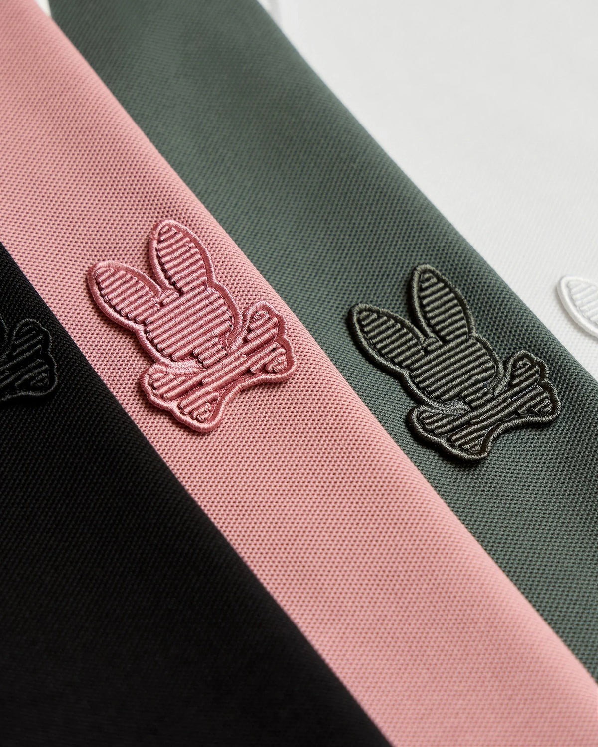 Close-up of the fabric from the MENS KINGSTON PIQUE POLO SHIRT - B6K764D200 by Psycho Bunny, showcasing embroidered bunny patches. The logos, featuring a bunny head with crossed bones underneath, are stitched in pink, black, and white on various Pima cotton strips in black, pink, and green.
