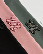 Close-up of the fabric from the MENS KINGSTON PIQUE POLO SHIRT - B6K764D200 by Psycho Bunny, showcasing embroidered bunny patches. The logos, featuring a bunny head with crossed bones underneath, are stitched in pink, black, and white on various Pima cotton strips in black, pink, and green.