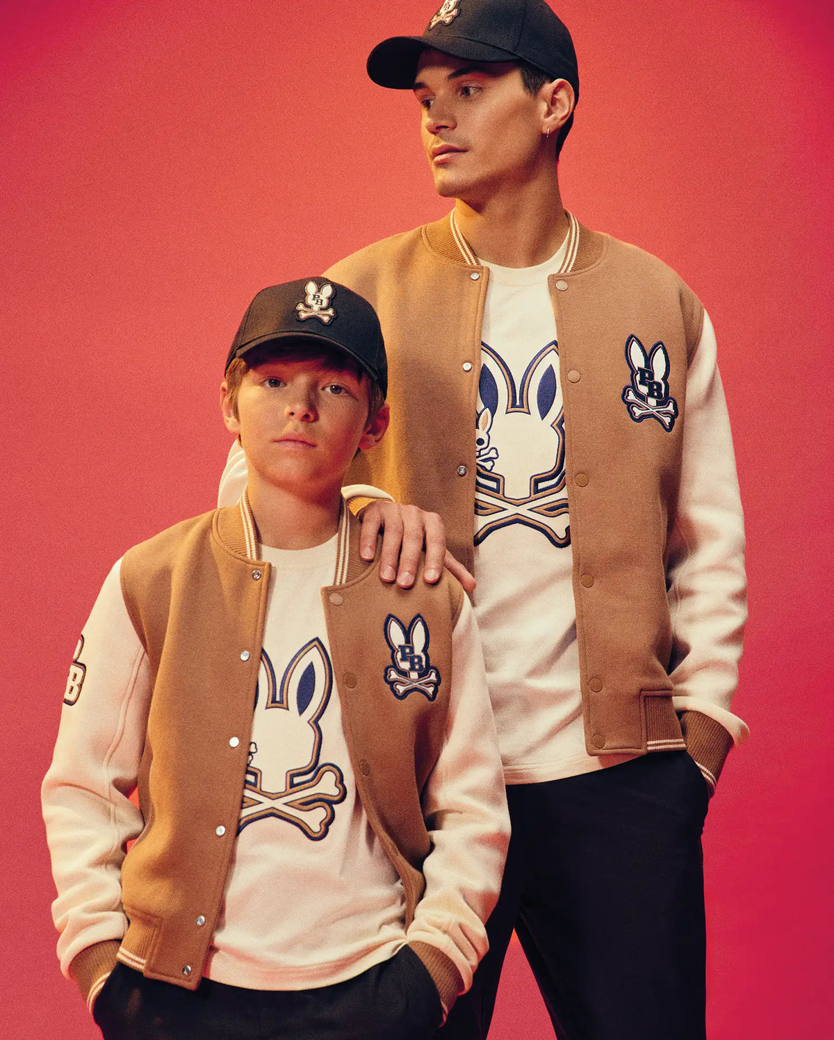 Two people pose against a pink background in matching tan and white Psycho Bunny KIDS WADE KNIT BOMBER jackets (KO1100001), with cartoon bunny logo shirts and black caps. The older rests a hand on the younger’s shoulder. Made from a premium cotton blend.