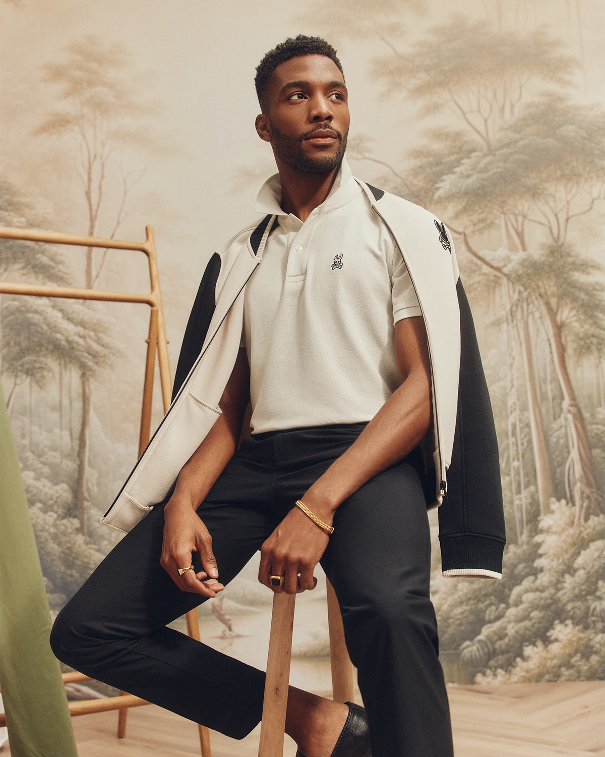Seated on a wooden stool, a person wears a Psycho Bunny MENS CLASSIC PIQUE POLO SHIRT (B6K001ARPC), layered with a black jacket, black pants, and shoes. They pose elegantly against serene forest scenery.