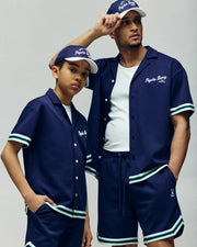 Two people pose confidently in matching Psycho Bunny KIDS BASIL TAPED SHORT - KB0900003, styled with navy baseball shirts, white undershirts, and navy caps against a neutral backdrop.