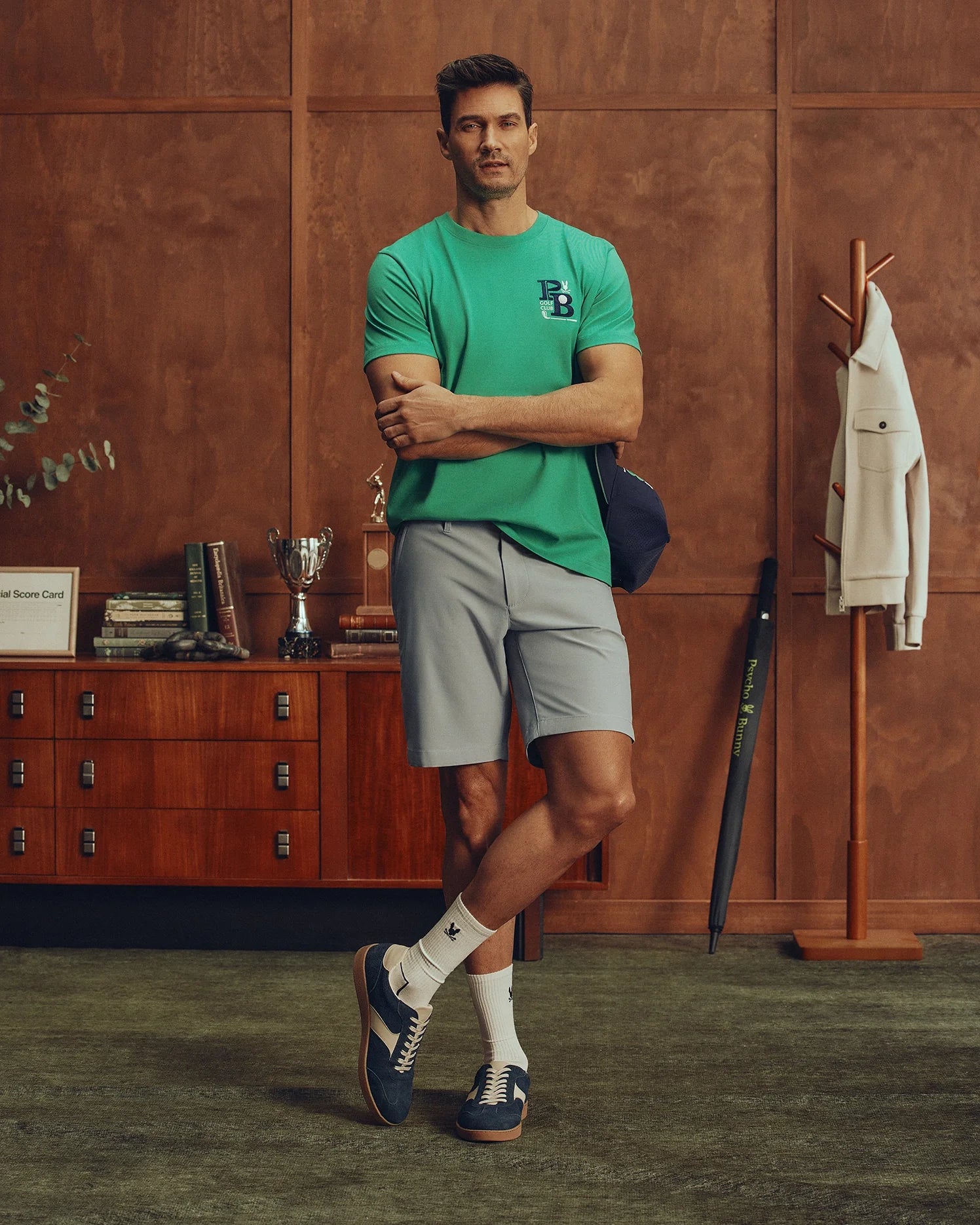A man in a Polo-inspired green MENS DENIS GRAPHIC TEE by Psycho Bunny and gray shorts stands cross-legged in a wood-paneled room. A wooden dresser displays books and a trophy behind him, while a coat rack on the right holds a white jacket and umbrella. Blue sneakers with logos complement his white socks.