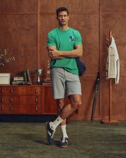 A man in a Polo-inspired green MENS DENIS GRAPHIC TEE by Psycho Bunny and gray shorts stands cross-legged in a wood-paneled room. A wooden dresser displays books and a trophy behind him, while a coat rack on the right holds a white jacket and umbrella. Blue sneakers with logos complement his white socks.