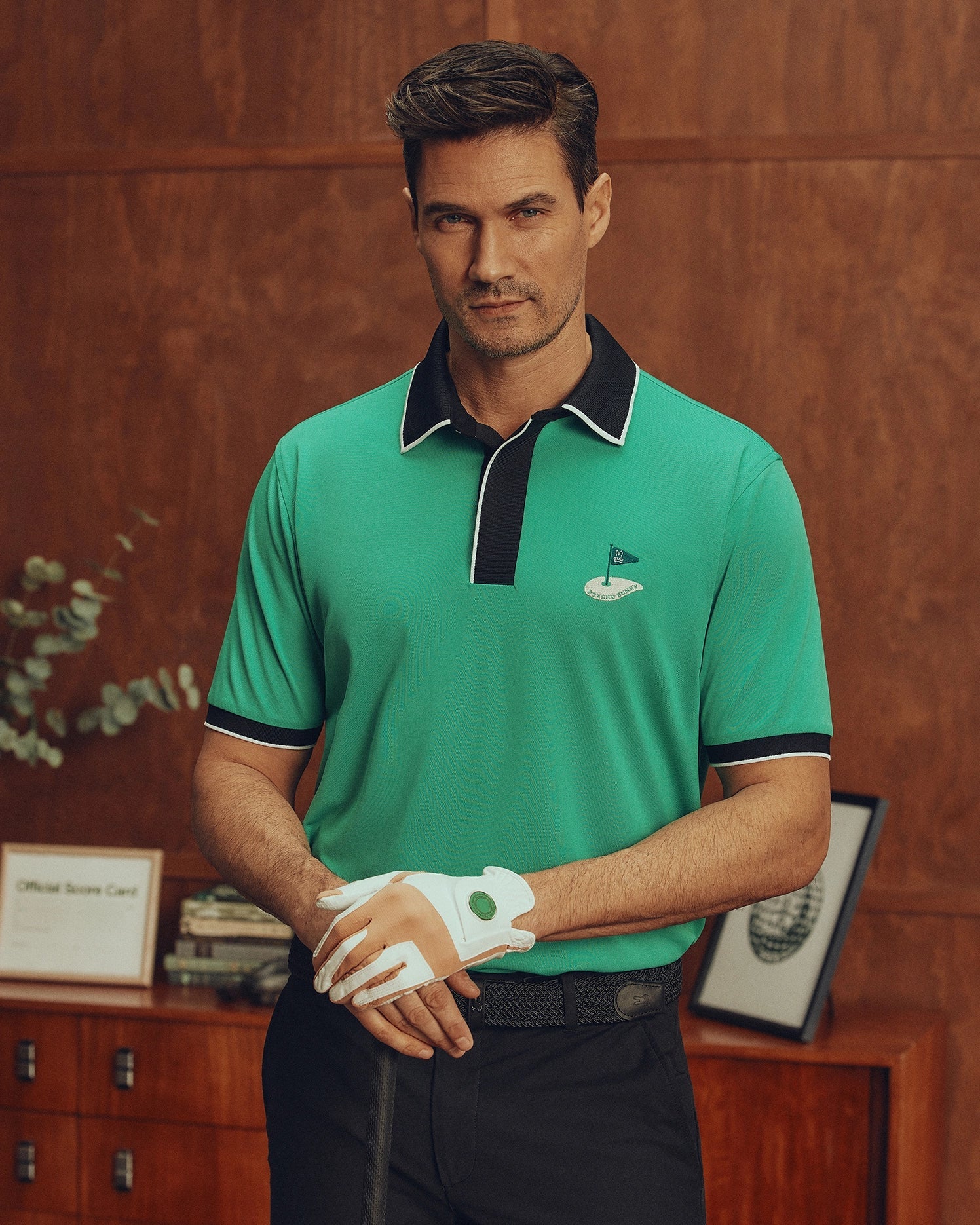 A man with dark hair stands indoors wearing a green Psycho Bunny MENS CLAYTON PIQUE POLO - MT0100004 made from breathable Pima cotton, complemented by a white glove as he holds a golf club. A brown wooden backdrop with books and framed items is visible behind him.