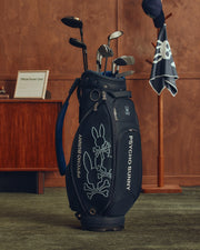 The OUTLINE BUNNY GOLF TOUR BAG - PBMG4FC1 by Psycho Bunny features a stylized rabbit and crossbones design on durable polyurethane, with "Psycho Bunny" text. It includes multiple clubs and a detachable strap. A wooden cabinet, scorecard, stand with towel, and hat are seen in the background.