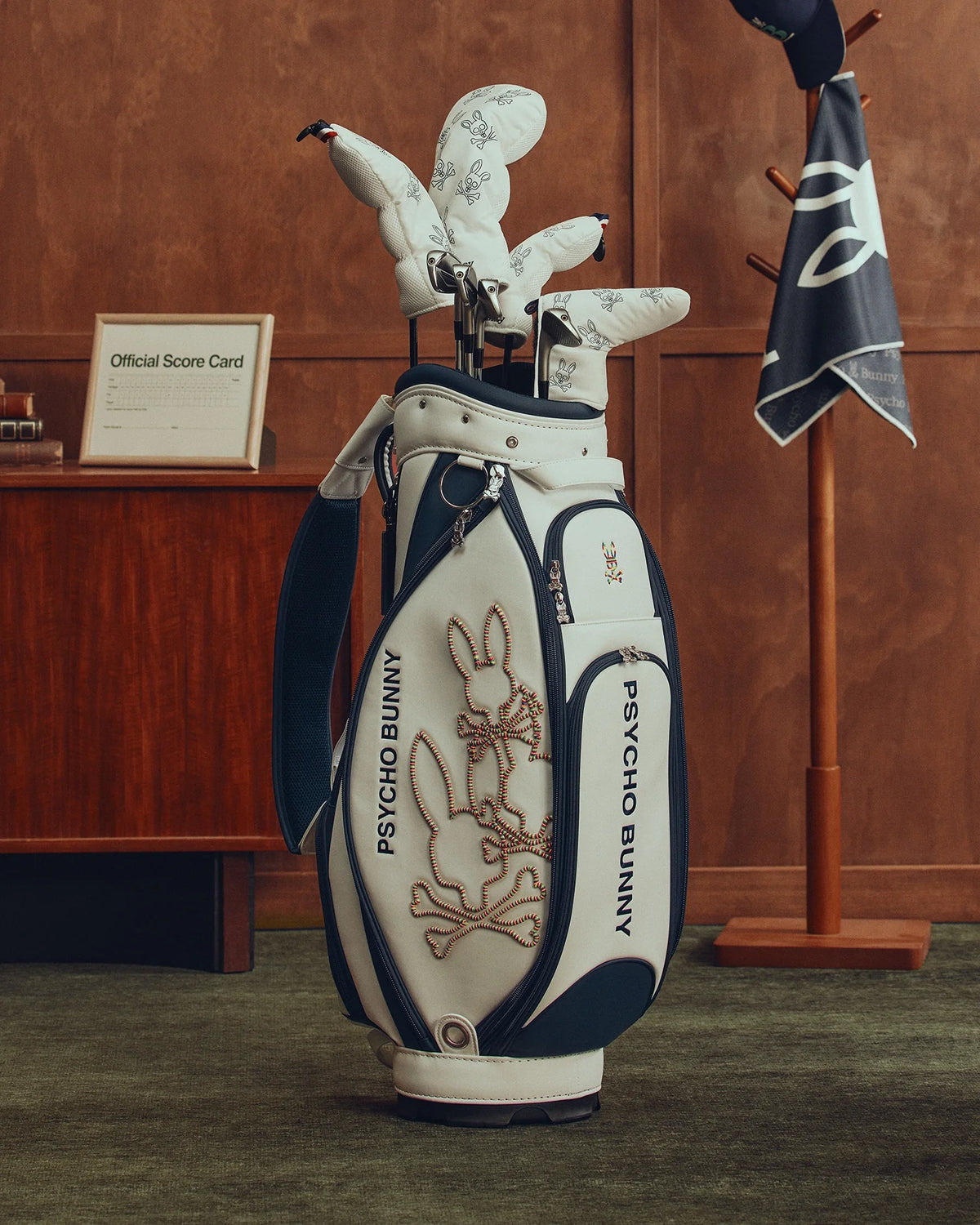 A stylish OUTLINE BUNNY GOLF TOUR BAG - PBMG4FC1 by Psycho Bunny rests on a green carpet in a wood-paneled room. Featuring bunny logos, it holds white golf club head covers. A stand with a hat and five-pocket dividers is visible in the background.