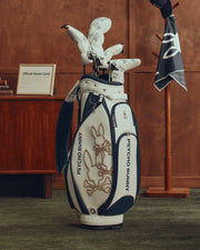 A stylish OUTLINE BUNNY GOLF TOUR BAG - PBMG4FC1 by Psycho Bunny rests on a green carpet in a wood-paneled room. Featuring bunny logos, it holds white golf club head covers. A stand with a hat and five-pocket dividers is visible in the background.