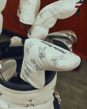 A golf bag stands indoors, showcasing putter covers adorned with a skull and crossbones design with bunny ears. Each white cover, marked "Psycho Bunny," adds edgy style to any golfer's collection. Enjoy the distinction of the MULTI BUNNY BLADE PUTTER COVER - PBMG4SH5 by Psycho Bunny.