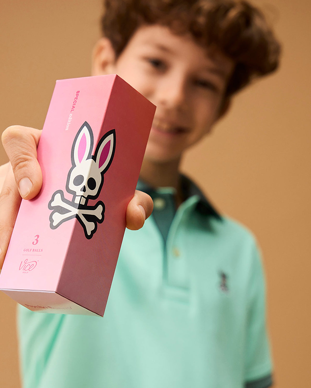 A boy with curly hair wearing a light green polo shirt is holding out a pink golf ball sleeve with a skull and crossbones design, where the skull has bunny ears. The box reads "SPECIAL EDITION" and "3 PACK GOLF BALLS - B6A565C200," featuring the Psycho Bunny logo to help minimize driver spin.