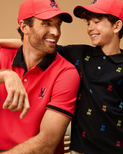 A man and a boy, both wearing red baseball caps, are smiling at each other. The man is dressed in a MENS KAYDEN PIQUE POLO - B6K675C200 by Psycho Bunny with a rabbit logo, and the boy wears a black Kayden pique polo shirt with colorful rabbit logos. The background is a plain, warm-toned wall.