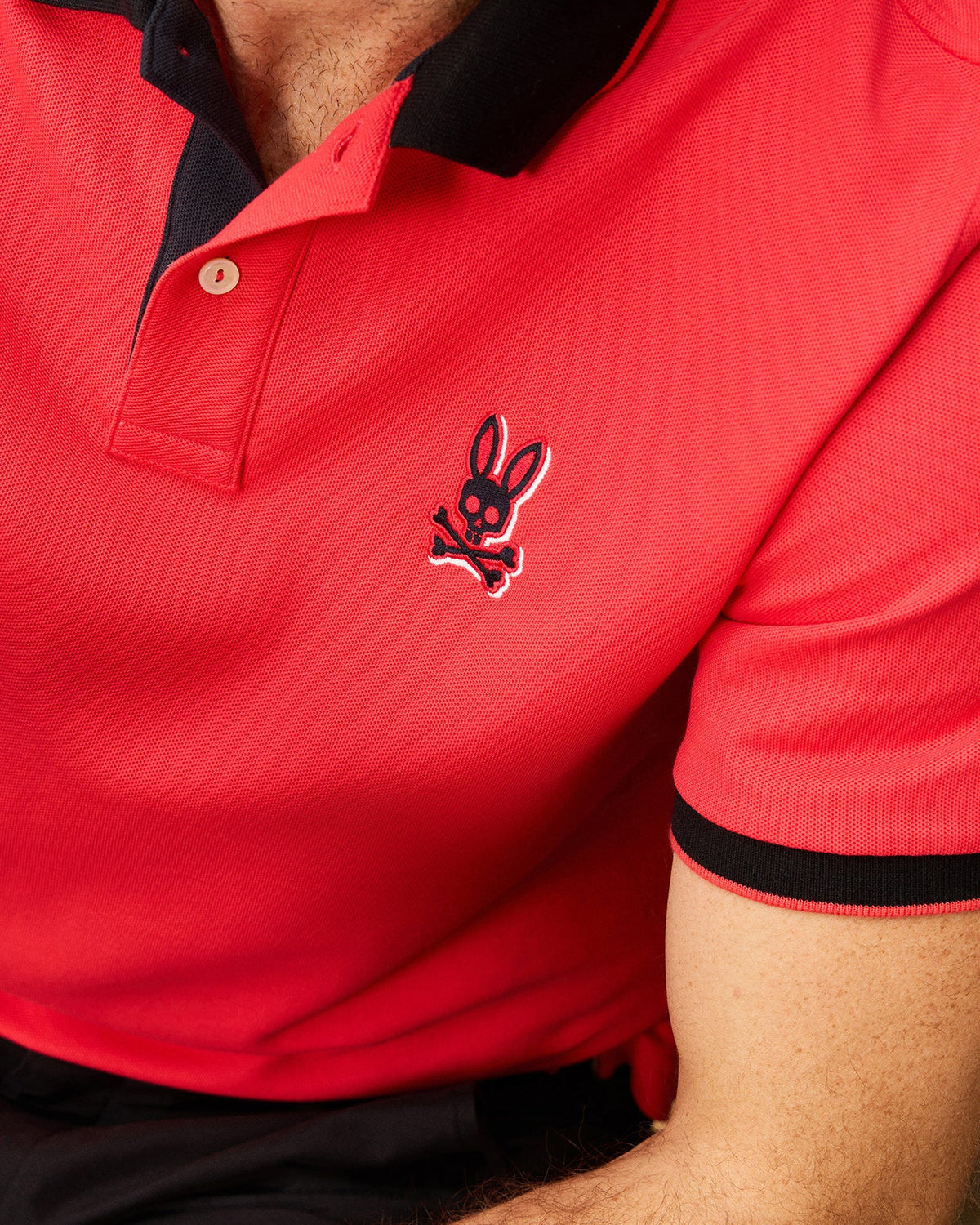 Close-up of a person wearing a red MENS KAYDEN PIQUE POLO - B6K675C200 from Psycho Bunny with black collar and sleeve trim. The shirt, made from diamond-knit Pima cotton, features an embroidered logo of a bunny with crossbones on the left chest. Only the upper torso and part of the arm are visible against a neutral background.