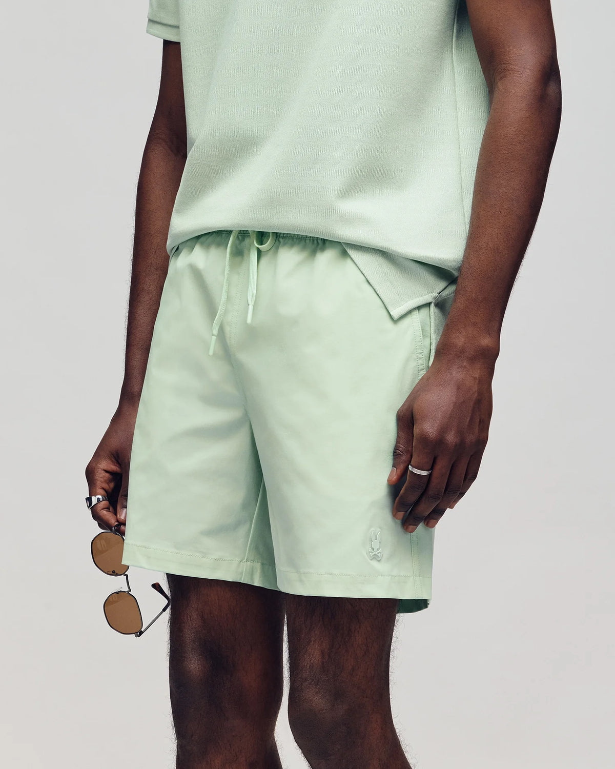 A person with medium skin tone stands in a light-colored setting, dressed in a light green T-shirt and the MENS SAMOS HYDROCHROMIC PRINTED SWIM TRUNK by Psycho Bunny, holding sunglasses. The monochromatic vibe is accentuated by the bunny patterns on their shorts.