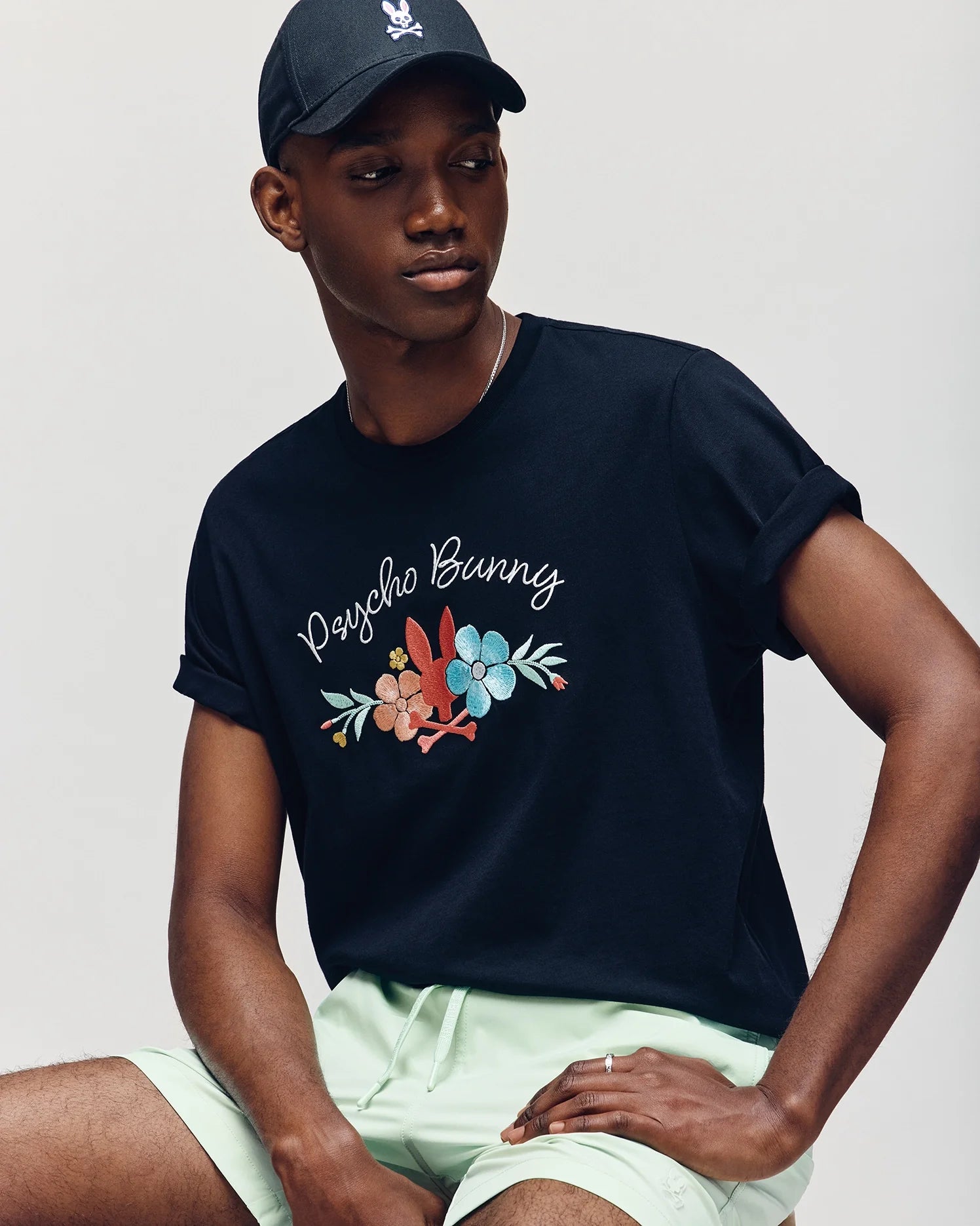 A man is wearing the Psycho Bunny MENS MEYLON GRAPHIC TEE - B6U656F200 in black with vibrant Bunny graphics, paired with light green shorts and a black cap, seated against a neutral background.