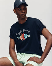 A man is wearing the Psycho Bunny MENS MEYLON GRAPHIC TEE - B6U656F200 in black with vibrant Bunny graphics, paired with light green shorts and a black cap, seated against a neutral background.