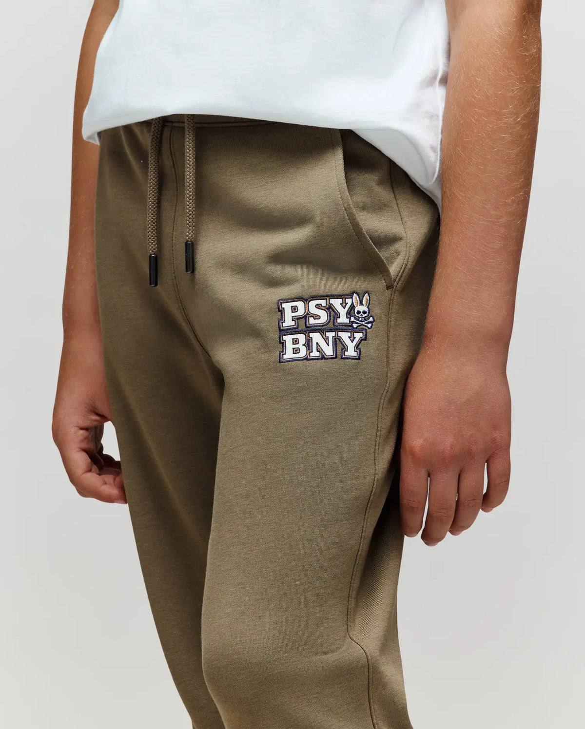 A child wearing Psycho Bunny KIDS WADE SWEATPANT - KB0800002 in olive green, featuring the "PSY BNY" logo and bunny graphic on the left thigh, made from a premium cotton blend, paired with a white t-shirt. Only torso and upper legs visible.