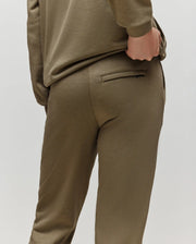 A child wears the Psycho Bunny KIDS WADE SWEATPANT (KB0800002) in olive green, premium cotton blend, posing from behind with one hand on the waistband near a welt pocket, set against a plain light background.