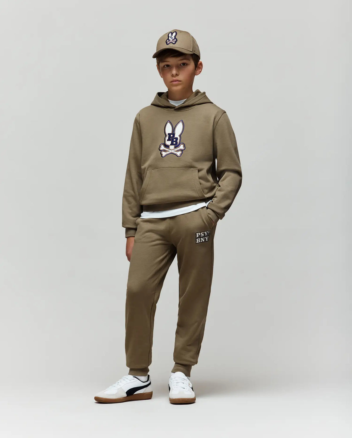 A young boy faces forward against a plain background, wearing Psycho Bunny’s KIDS WADE SWEATPANT (KB0800002) in tan with jogger pants, a hoodie featuring the cartoon bunny logo, a matching cap, and white sneakers. His hands are in his pockets.