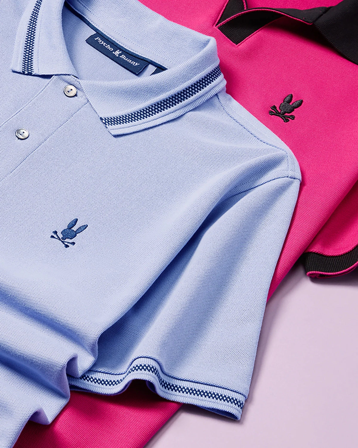 Two folded MENS RICHARDS JOHNNY COLLAR PIQUE POLO SHIRTS by Psycho Bunny, one in light blue and the other in hot pink, are displayed on a flat surface. Both breathable Pima cotton polos feature an embroidered black bunny logo on the chest. The light blue shirt has a checkered collar and sleeve trim, while the hot pink shirt sports black accents.