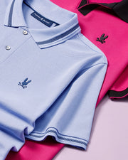 Two folded MENS RICHARDS JOHNNY COLLAR PIQUE POLO SHIRTS by Psycho Bunny, one in light blue and the other in hot pink, are displayed on a flat surface. Both breathable Pima cotton polos feature an embroidered black bunny logo on the chest. The light blue shirt has a checkered collar and sleeve trim, while the hot pink shirt sports black accents.