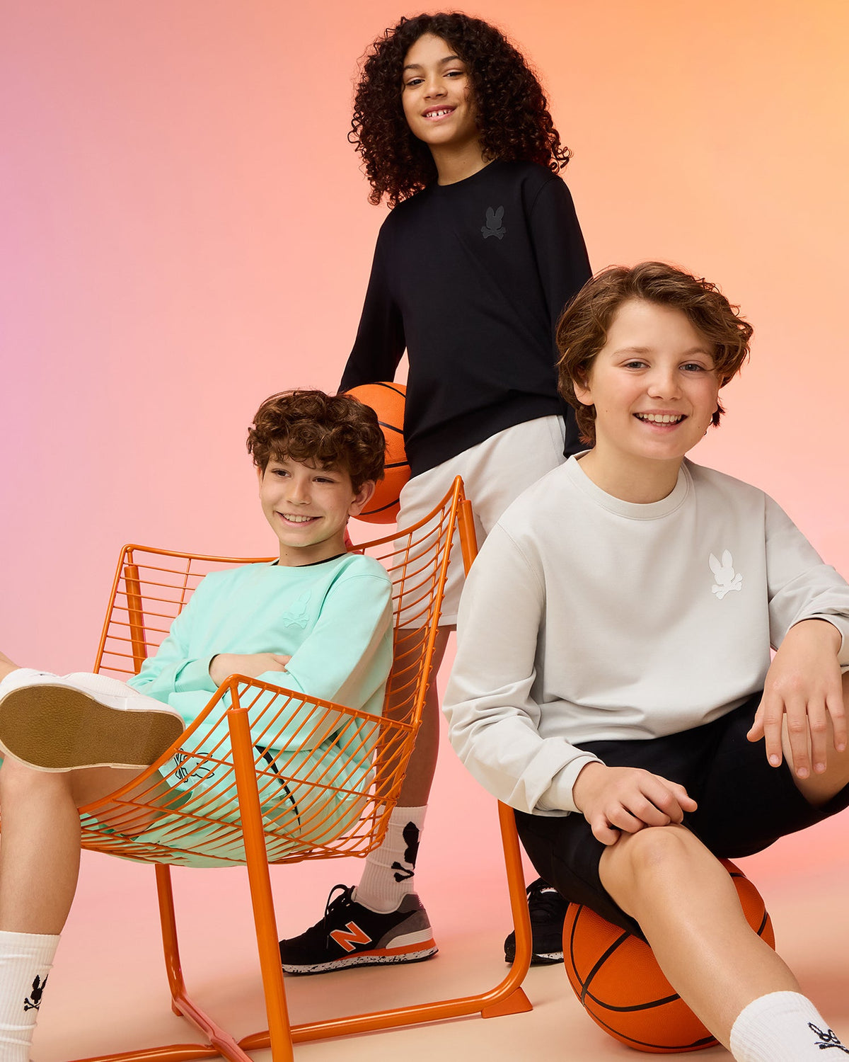 Three children pose with basketballs against a gradient background. One sits on an orange chair, sporting a mint green outfit made from premium cotton fabric. The others stand, dressed in the KIDS HOUSTON FRENCH TERRY CREWNECK by Psycho Bunny in black and light gray, all smiling in their casual sneakers.