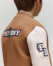 A child wears the Psycho Bunny KIDS WADE KNIT BOMBER (KO1100001), a brown and white varsity jacket with “PSYBNY” and a bunny logo on the back, plus a “PB” patch with a small bunny head on the sleeve. Seen from behind.